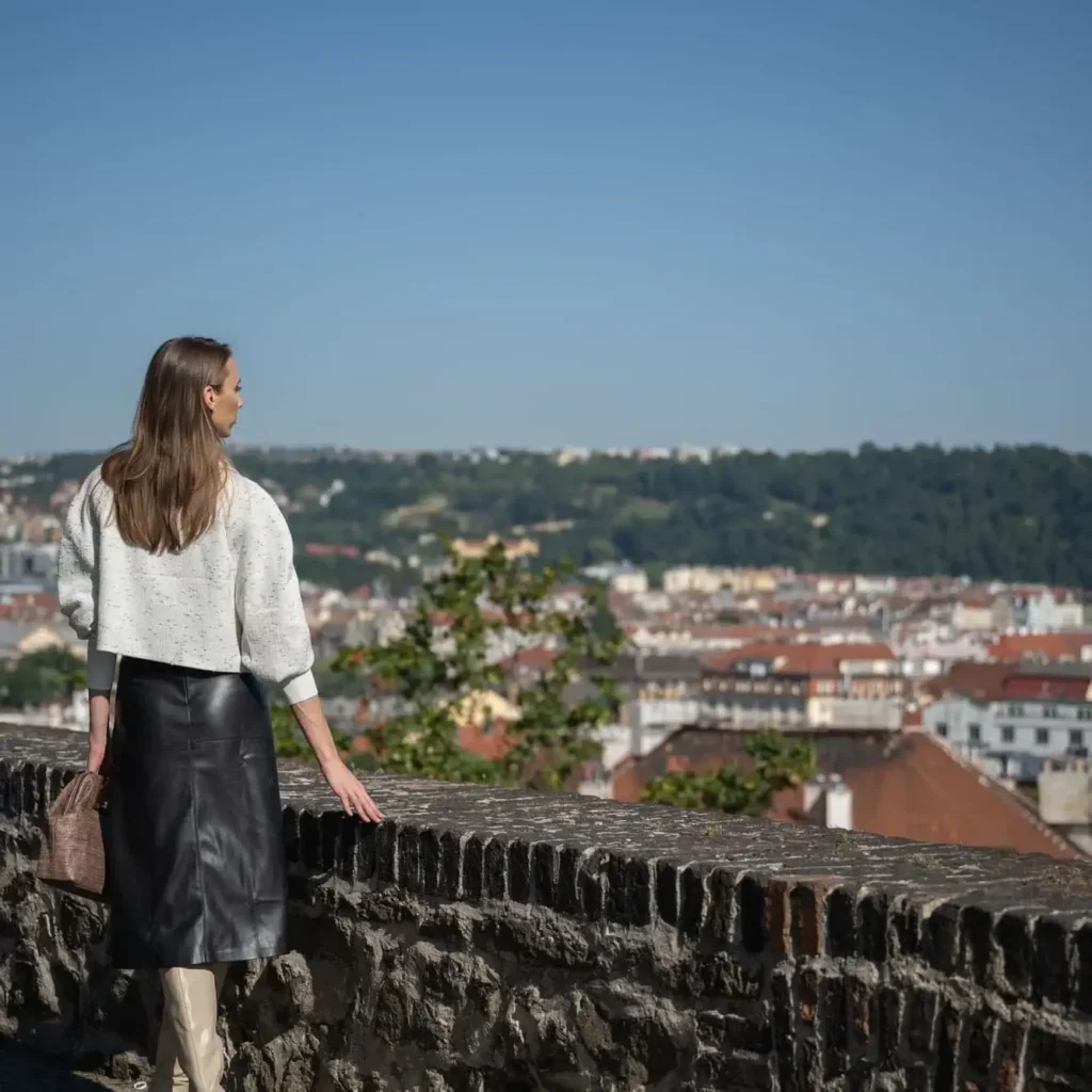 A woman in a white top and black skirt stands by a stone wall during one of our Signature Tours, overlooking a cityscape with red-roofed buildings and green hills beneath a clear blue sky.